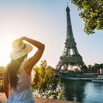 Touristin mit Sonnenhut und weißem Kleid steht bei Sonnenuntergang vor dem Eiffelturm in Paris