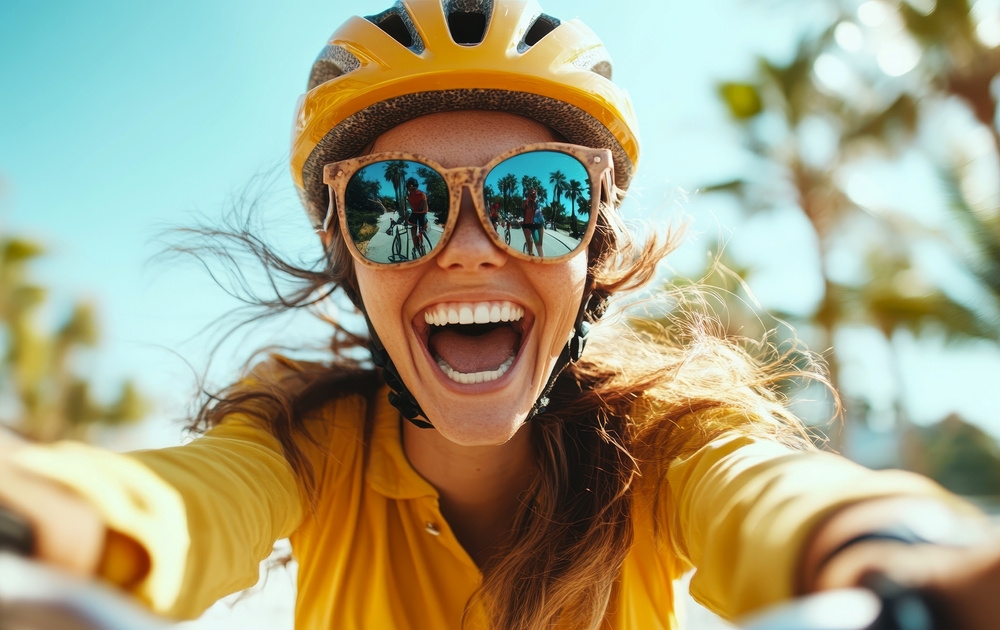  junge Frau, die mit einem gelben Helm und einer stylischen Sonnenbrille Fahrrad fährt, umgeben von Palmen an einem sonnigen Tag