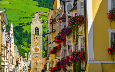 Straße mit historischen Gebäuden und Uhrturm, Balkonblumen, grüne Hügel im Hintergrund.