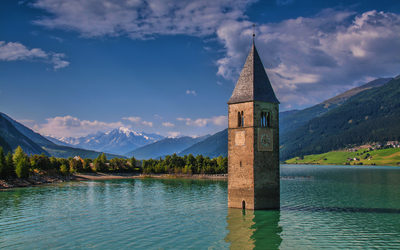 Kirchturm eines versunkenen Dorfes im Reschensee, umgeben von Bergen und blauem Himmel.