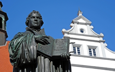 Statue eines Mannes mit aufgeschlagenem Buch vor blauem Himmel und historischem Gebäude.