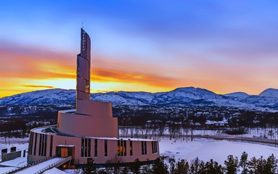 Eine moderne Kirche im Schnee vor einer Bergkulisse während eines bunten Sonnenuntergangs.