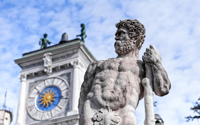 Steinstatue eines bärtigen Mannes vor einem Gebäude mit Uhr und Figuren.
