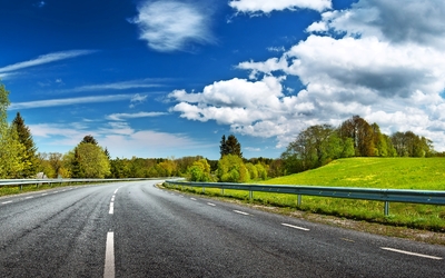 Landstraße mit Kurve, umgeben von grünen Feldern und Bäumen, unter blauem Himmel.