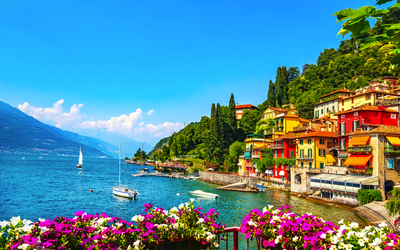 Bunte Häuser am Seeufer mit Blumen im Vordergrund und Segelbooten im Wasser.