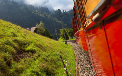 Roter Zug fährt durch grüne Berglandschaft bei sonnigem Wetter.