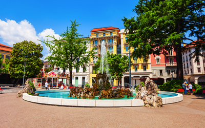 Springbrunnen auf einem Stadtplatz mit bunten Gebäuden im Hintergrund.