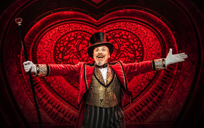 Mann in rotem Frack, Zylinder und mit Gehstock auf einer Bühne vor rotem Hintergrund.