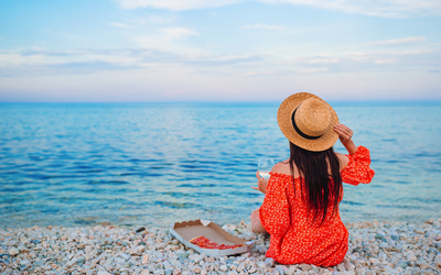 Frau am Kiesstrand mit Strohhut, schaut aufs Meer, hält ein Glas, Pizzaschachtel neben ihr.