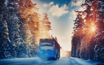Verschwommener Bus auf schneebedeckter Straße durch winterlichen Wald bei Sonnenuntergang.