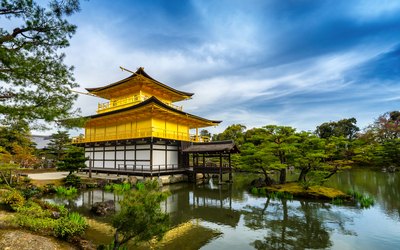 Ein goldener Tempel in Japan am Wasser, umgeben von Bäumen und blauem Himmel.