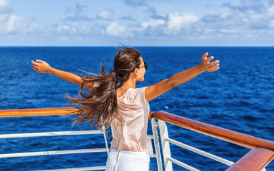 Frau auf einem Schiffsdeck blickt aufs Meer mit ausgebreiteten Armen.